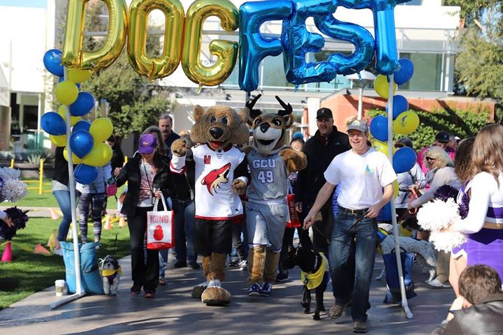 A Group of People and Dogs Walk With Two Dog Mascots During a Leap Day DogFest Event.