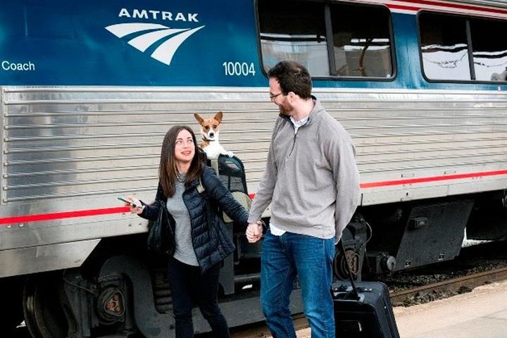 Two people and a dog in a backpack in front of Amtrak train.